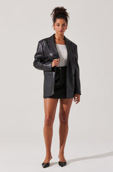 A woman is wearing a black leather blazer with a notched lapel collar, paired with a sleeveless white top with a textured pattern, a black mini skirt, and black pointed-toe heels with bow details.